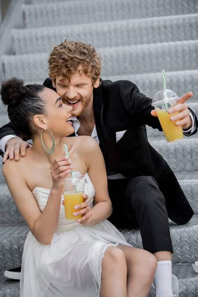 cheerful redhead groom pointing with finger near african american bride with orange juice on street - Zdjęcie, obraz