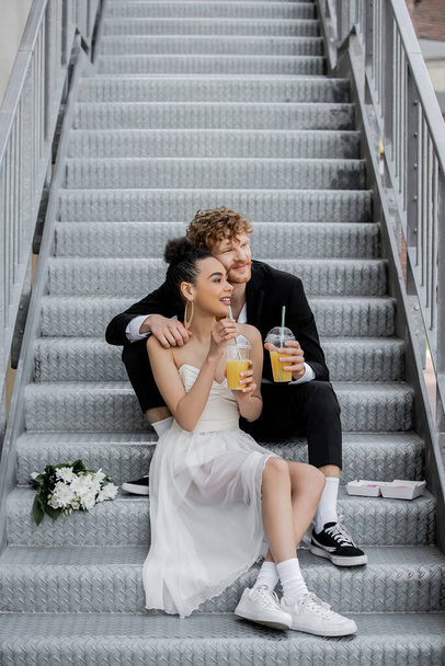 happy interracial newlyweds with orange juice looking away on street stairs near bouquet - Foto, afbeelding