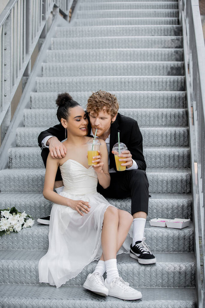 redhead man drinking orange juice from straw near african american bride, sitting on stairs in city - 写真・画像