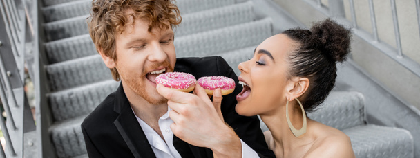 wedding, urban setting, fun, interracial newlywed couple feeding each other with donuts, banner - Foto, afbeelding