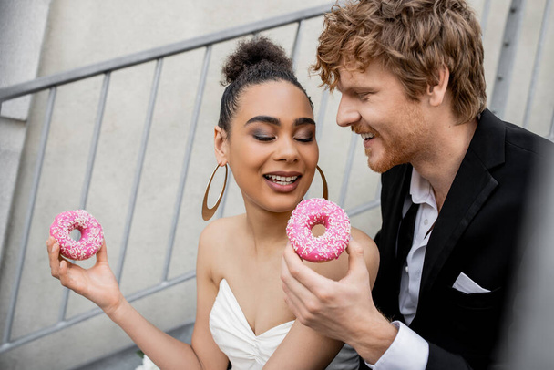 радісна багатоетнічна пара у весільному вбранні, що тримає солодкі пончики, весілля в міській обстановці - Фото, зображення