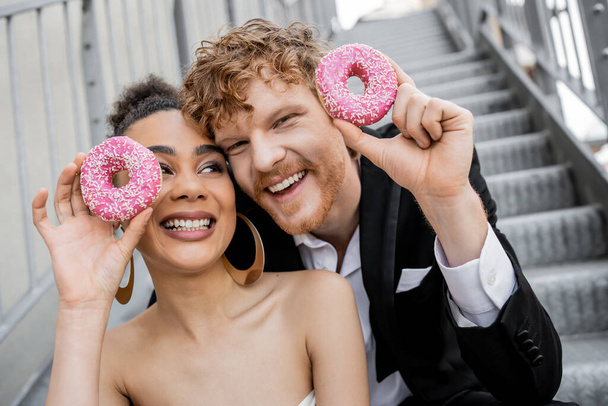 delighted and elegant multiethnic newlyweds posing with sweet donuts, wedding in urban setting - Foto, afbeelding