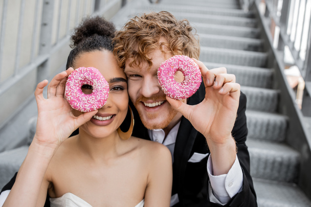having fun, wedding in city, excited interracial newlyweds obscuring face with donuts - Photo, Image