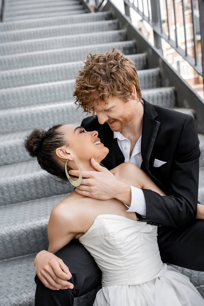 smiling redhead man hugging young african american bride on stairs in city, tenderness, happiness - Фото, изображение
