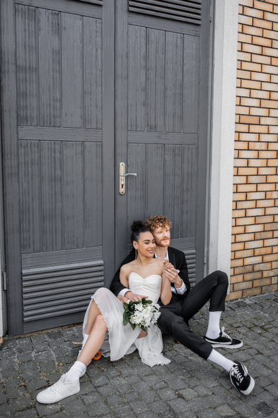 feliz casal interracial sentado no pavimento da rua perto de portas, traje de casamento, flores - Foto, Imagem