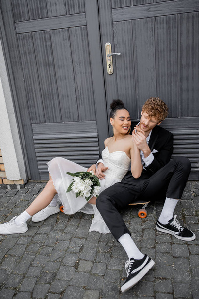 redhead man holding hands of african american bride with flowers, sitting on pavement near doors - Fotografie, Obrázek