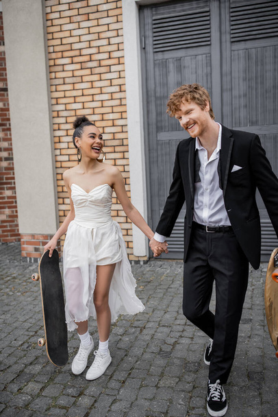 cheerful interracial newlyweds walking with longboard and skateboard on city street, wedding attire - Photo, Image