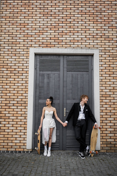 carefree multiethnic newlyweds with longboard and skateboard holding hands near city building - 写真・画像