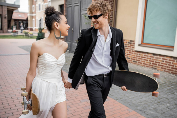 multiethnic couple walking with longboard and skateboard on street, wedding outfit, sunglasses - Foto, afbeelding