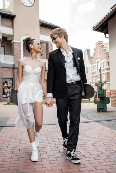 wedding in city, romantic interracial couple walking with longboard and skateboard on street - Photo, Image