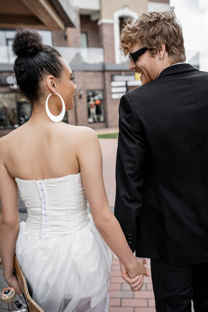 redhead groom in sunglasses, african american bride with longboard looking at each other on street - Photo, Image