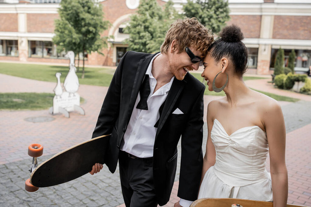 overjoyed multiethnic newlyweds with longboard and skateboard on street, sunglasses, wedding outfit - Photo, Image