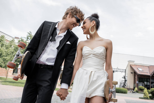 ロングボードとスケートボードの若い異人種間の新婚夫婦は,通りで手を保持し,幸福 - 写真・画像