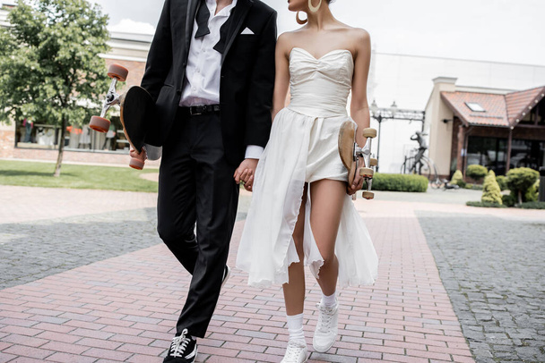 vista ritagliata di elegante coppia multietnica passeggiando con longboard e skateboard, matrimonio in città - Foto, immagini