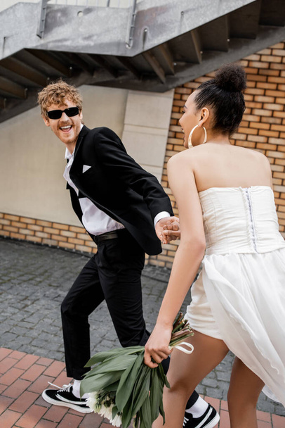 marié excité dans des lunettes de soleil tenant la main de la mariée afro-américaine et courant sur la rue urbaine - Photo, image
