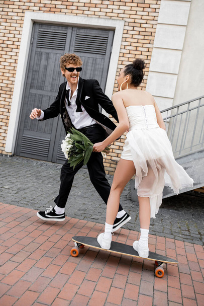 excited groom in sunglasses near african american bride in wedding dress riding longboard - Zdjęcie, obraz