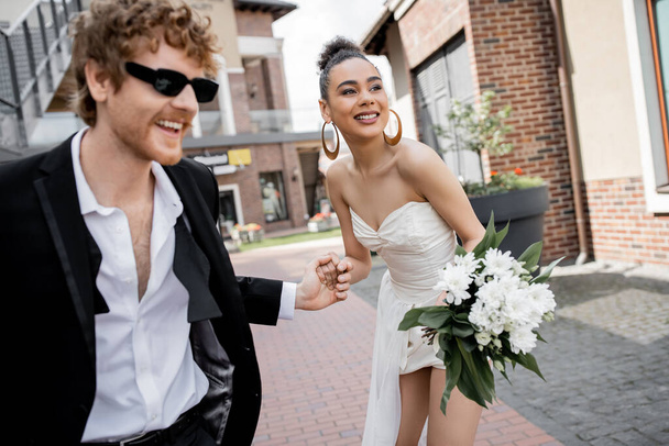 afroamericana americana novia con flores y pelirroja novio en gafas de sol de la mano en la calle - Foto, imagen