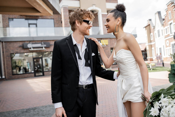 outdoor wedding, modern and stylish interracial newlyweds looking at each other on street - Foto, imagen