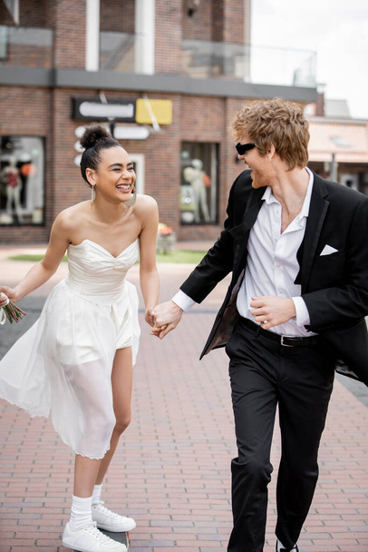 romance urbano, elegante interracial recién casados tomados de la mano y corriendo en la ciudad europea - Foto, Imagen