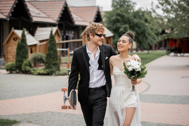 χαρούμενο διαφυλετικό ζευγάρι περπατά στην πόλη με longboard και λουλούδια, γαμήλια ενδυμασία, γυαλιά ηλίου - Φωτογραφία, εικόνα