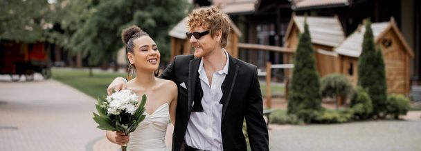 matrimonio all'aperto, sposa afroamericana con fiori e sposo in occhiali da sole passeggiando in città, striscione - Foto, immagini