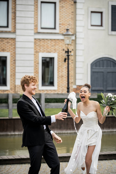 sposa afro-americana eccitata vicino allo sposo con champagne versato dalla bottiglia, celebrazione all'aperto - Foto, immagini