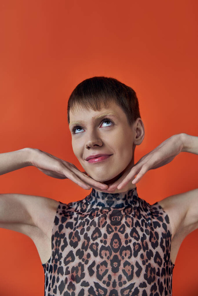 happy androgynous person smiling and posing on orange backdrop, hands near face, queer fashion - Photo, Image