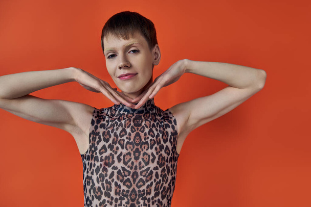 cheerful androgynous person smiling and posing on orange backdrop, hands near face, queer fashion - Photo, Image
