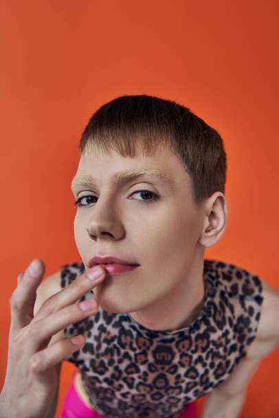 nonbinary person smiling and touching lips on orange backdrop, looking at camera, queer fashion - Photo, Image