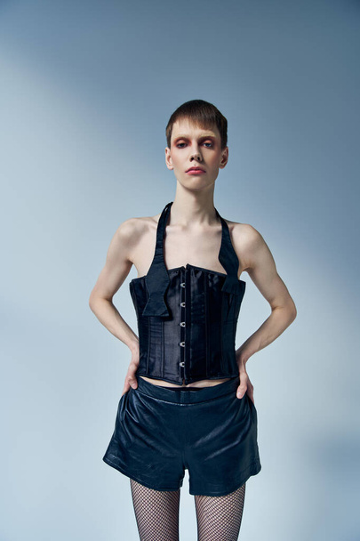 queer model in black corset and shorts posing with hands on hips on grey backdrop, androgynous - 写真・画像
