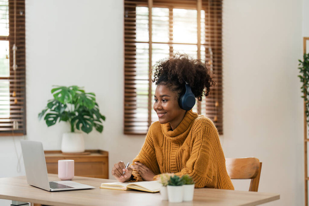 Enseignement à distance. Portrait d'une femme souriante afro-américaine assise au bureau, utilisant un ordinateur portable et écrivant dans un carnet, prenant des notes, regardant un tutoriel, une conférence ou un webinaire, étudiant en ligne à la maison. - Photo, image