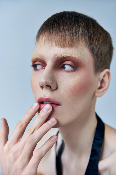 queer person with makeup looking away on grey backdrop, androgynous, touching lip, self expression - Foto, immagini
