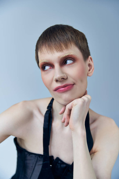 queer avec maquillage regardant loin sur gris, modèle androgyne, expression de soi, sceptique - Photo, image