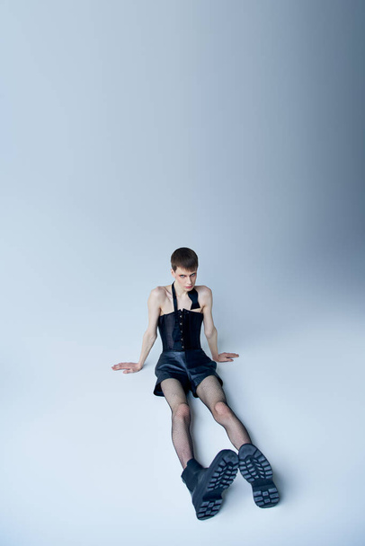 queer person in black corset and fishnet tights sitting on grey, androgynous model, lgbt fashion - Foto, Imagen