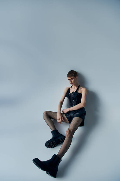 modelo andrógino en corsé, pantalones cortos y medias de rejilla posando sobre fondo gris, lgbt, moda queer - Foto, Imagen