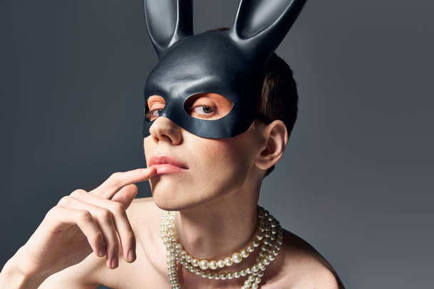 retrato, modelo queer en corsé y bdsm conejito máscara posando en gris, dedo cerca de los labios, moda - Foto, imagen