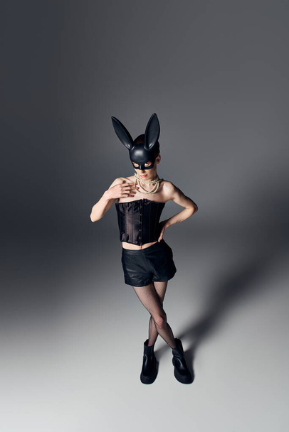 vet look, gender vloeistof persoon in korset poseren in bdsm bunny masker op grijs, queer stijl, hand op heup - Foto, afbeelding