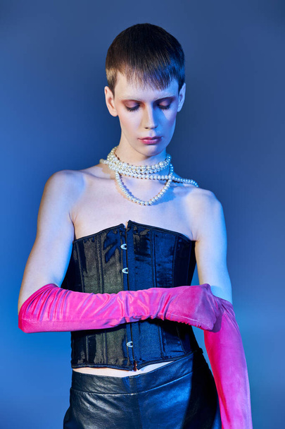 persona queer en corsé negro y collar de perlas con guantes de color rosa sobre fondo azul, estilo vanguardista - Foto, imagen