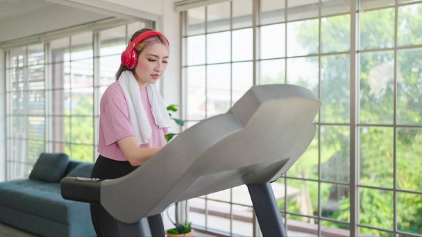 Vrolijke jonge Aziatische vrouwen in sportkleding dragen een koptelefoon tijdens het trainen op de machine thuis. Jonge vrouwen trainen op een elliptische machine. Gezondheidszorgconcept - Foto, afbeelding