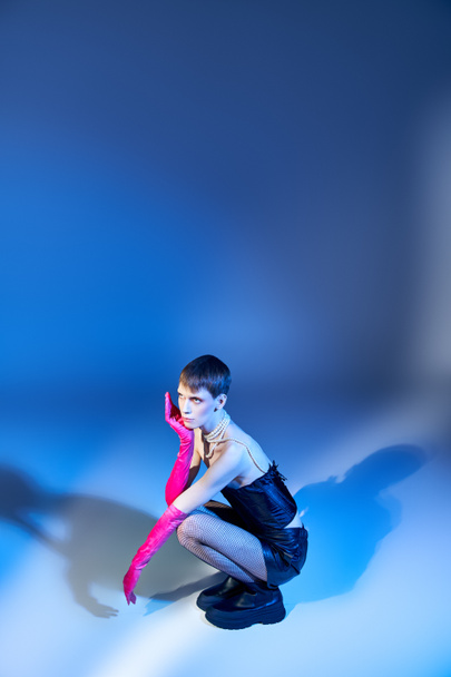 buzerant v korzetu a černých šortkách sedící na modrém pozadí, nebinární model v růžových rukavicích - Fotografie, Obrázek