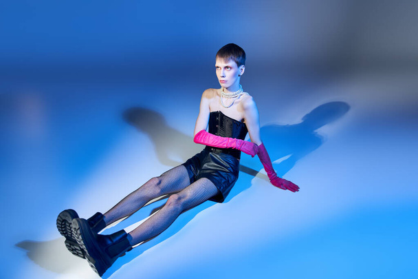 model in corset and boots sitting on blue backdrop, nonbinary person in pink gloves, queer fashion - Fotografie, Obrázek
