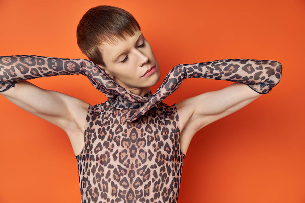modello queer in abito stampa animalier in posa su sfondo arancione, occhi chiusi, elegante genderfluid - Foto, immagini