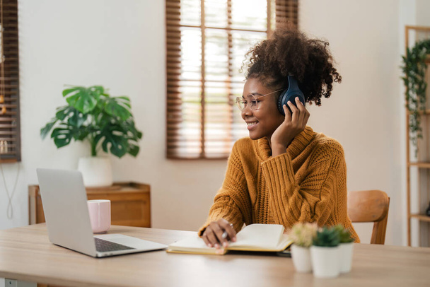 Lächelnde junge afrikanisch-amerikanische Frau mit drahtlosen Kopfhörern sitzt am Schreibtischtisch und arbeitet am Laptop und schreibt Briefe in Papiernotizbuch, macht Notizen, während sie Weninar beobachtet. - Foto, Bild