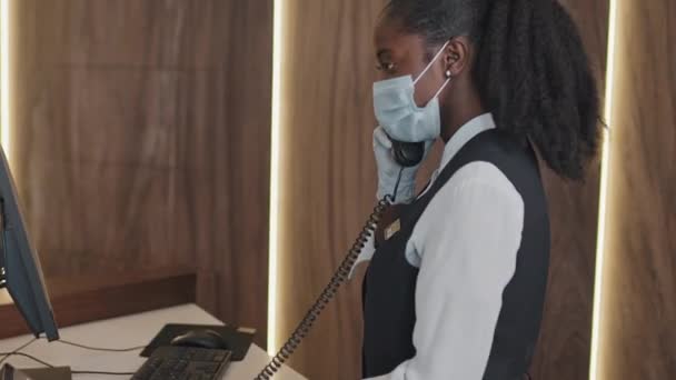 Молодая афроамериканка в защитной маске и перчатках, работающая на стойке регистрации, отвечает на телефонный звонок - Кадры, видео