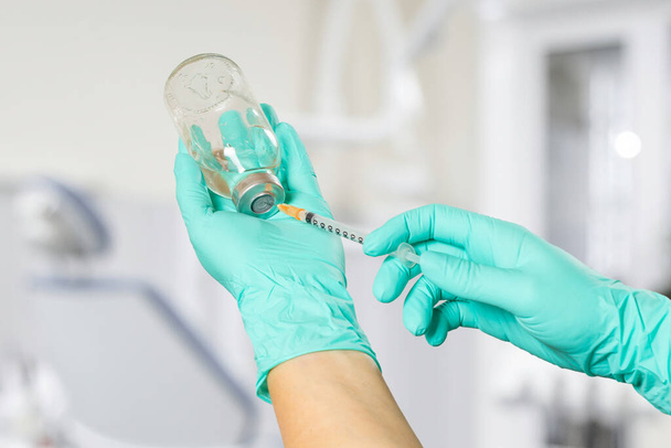 Glasflasche für flüssige Medizin und eine Plastikspritze für die Injektion in die Hände in Latex-Handschuhe. Zahnarztpraxis im Hintergrund. - Foto, Bild