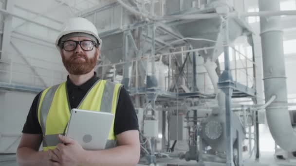 Confident mature Caucasian male engineer with beard on face wearing uniform with hardhat and eyeglasses holding digital tablet looking at camera - Footage, Video
