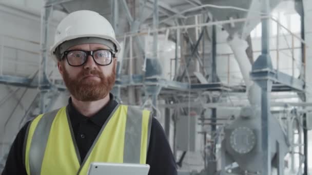 Medium close-up portrait of mature Caucasian man with beard on face wearing hardhat and eyeglasses working in modern plant looking at camera - Footage, Video