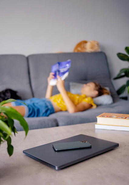 cyfrowy detox i rekreacji koncepcja - zbliżenie gadżetów w koszyku na stole i dziewczyny czytanie książki w domu - Zdjęcie, obraz