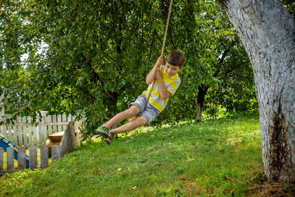 Fröhlicher kleiner Junge hat Spaß auf einer Seilschaukel, die er beim Ausruhen außerhalb der Stadt gefunden hat. Aktive Freizeit mit Kindern. - Foto, Bild