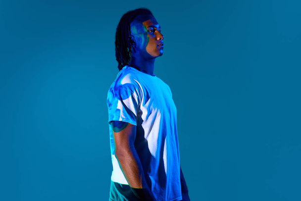 Young african man with neon light reflection on body standing against blue background in neon light. Innovations. Concept of art, fashion, modern style, cyberpunk, futurism, ad - Photo, Image